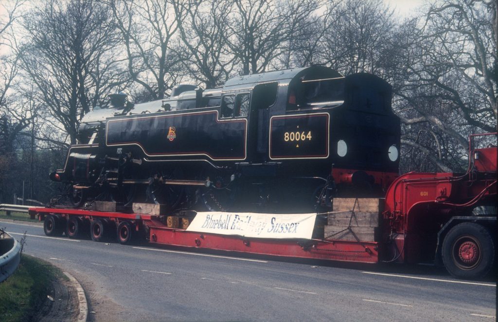 80064 loaded on a red road lorry beside a country road, with a Bluebell Railway Sussex banner on the trailer.