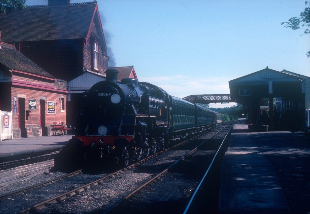 80064 at the platform of a red-brick station decorated with old enamel advertising signs, green coaches behind.