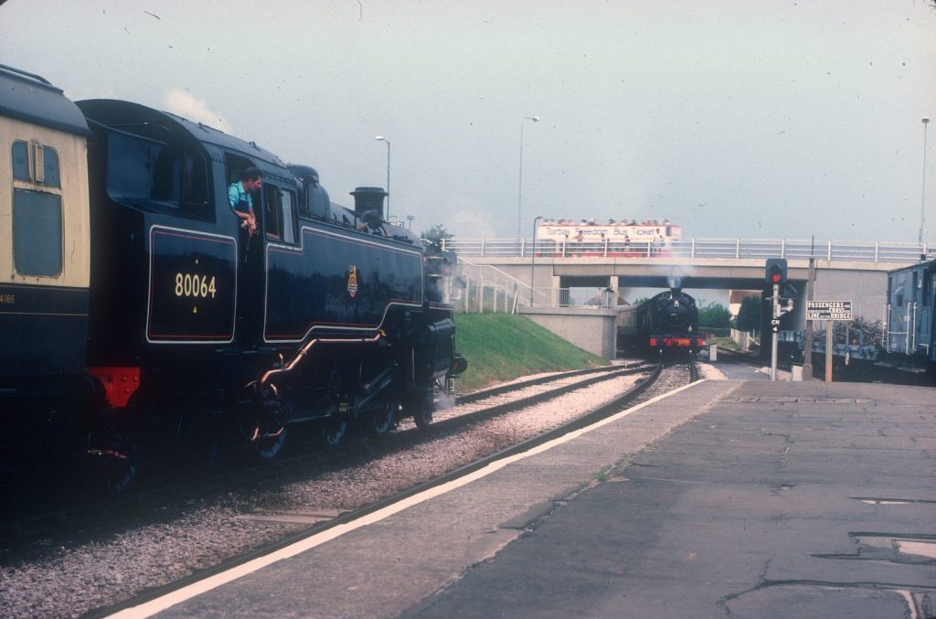 80064 in the foreground with its driver looking ahead, as 5239 approaches under a road bridge carrying an open-top bus.