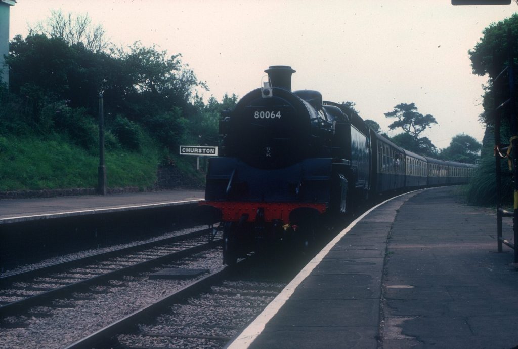 Front view of 80064 at Churston station platform with a long train of coaches curving behind it.