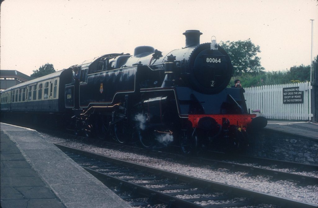 Three-quarter front view of 80064 standing at a station platform beside a white picket fence.