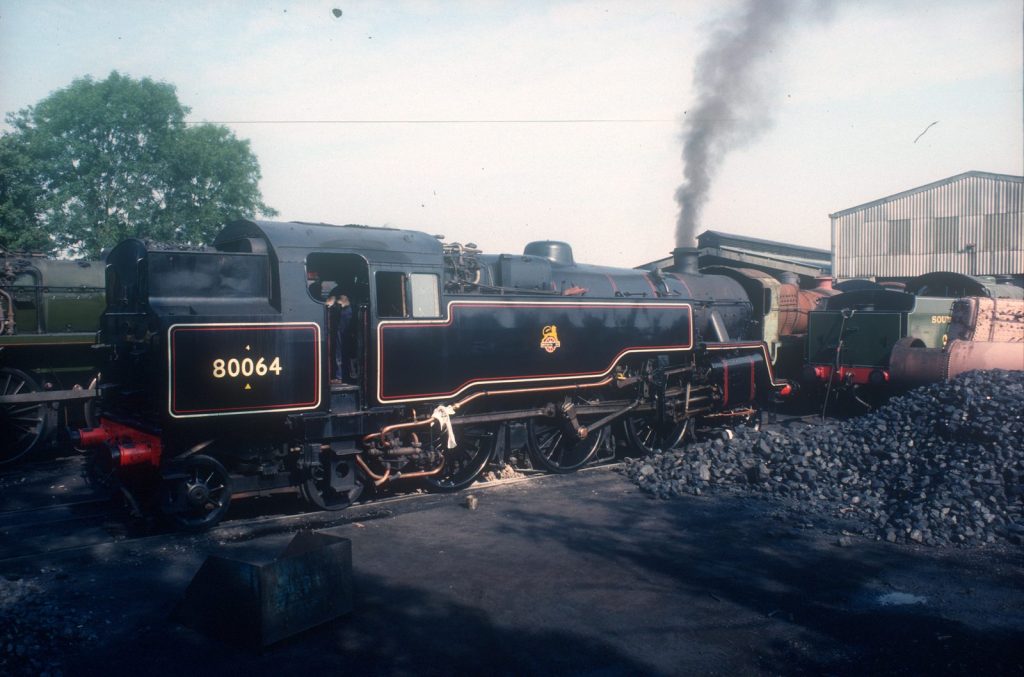 80064 standing by heaps of coal outside an engine shed, smoke rising from its chimney, other engines nearby.