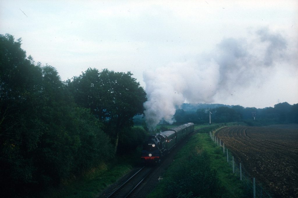 80064 in the distance leading a train past trees and a ploughed field, steam billowing above.