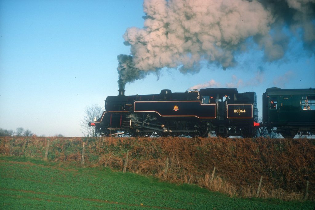 Side view of 80064 on an embankment under a blue sky, a tall plume of smoke and steam above, pulling a green coach.