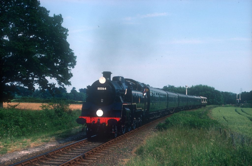 Front view of 80064 with lamps lit, pulling a long train of green coaches between fields.