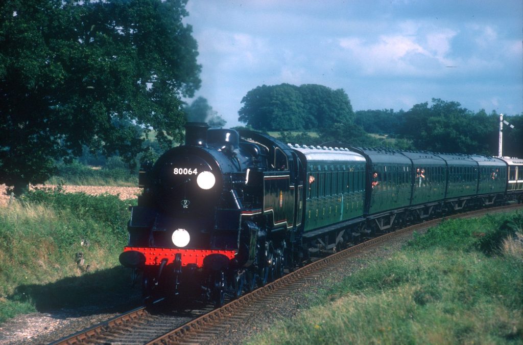 80064 head-on with two white lamps lit, leading green coaches round a curve on a sunny day.