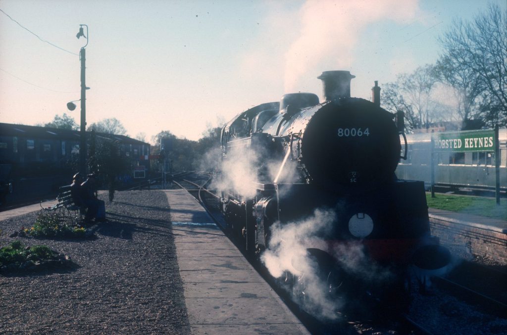 80064 at the platform at Horsted Keynes station, steam around its wheels and the green station nameboard on the right.