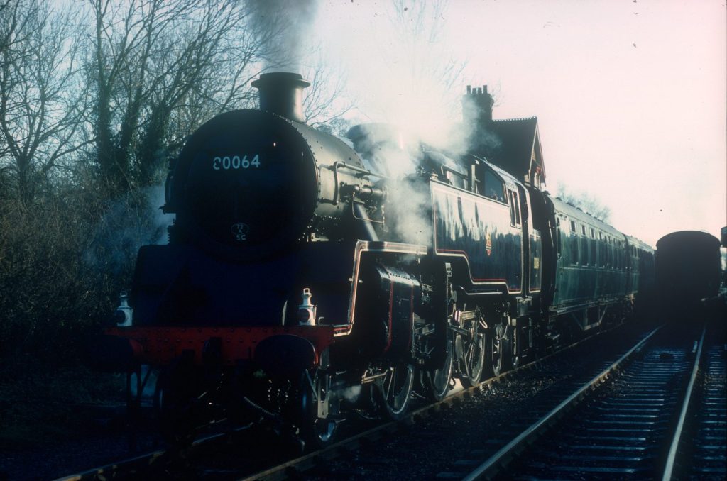 Three-quarter front view of 80064 at a station, steam drifting around it and coaches behind.