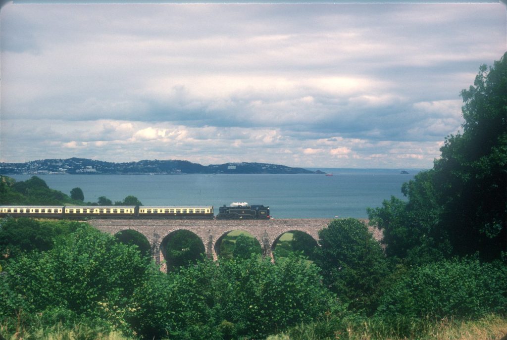 80064 hauling a long train of cream coaches across a stone viaduct, with a wide bay and a distant town beyond.