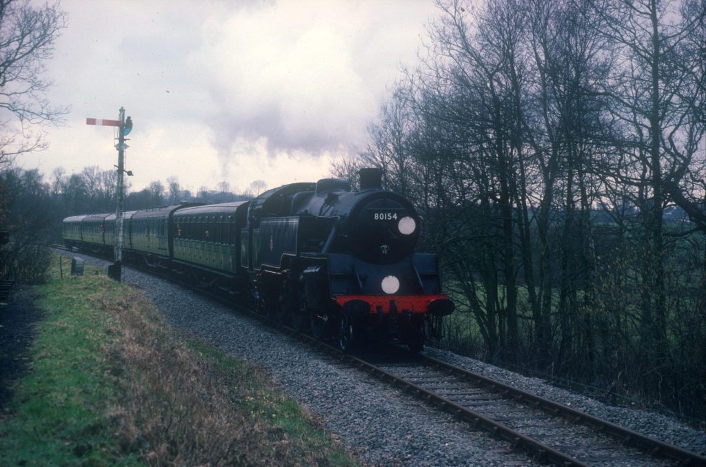 80064 with lamps lit, leading green coaches past a semaphore signal among bare winter trees.