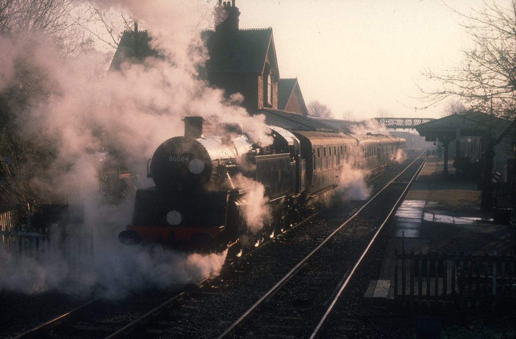 80064 at a station platform almost hidden by clouds of steam, the station building behind.